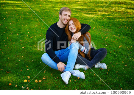 Happy young couple sit in an embrace in the park on the grass Happy young couple sit in an embrace in the park on the grass 100990969