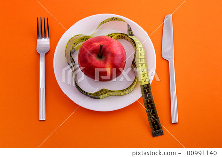 red apple on plate with measuring tape, knife and fork on an orange background red apple on plate with measuring tape, knife and fork on an orange background 100991140