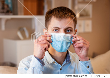 young guy sitting on the couch wearing a medical mask young guy sitting on the couch wearing a medical mask 100991178