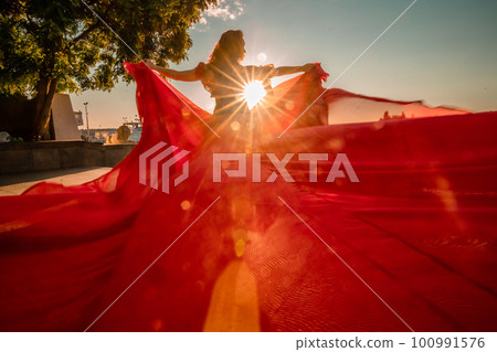 Sunrise red dress. A woman in a long red dress against the backdrop of sunrise, bright golden light of the sun's rays. The concept of femininity, harmony. 100991576
