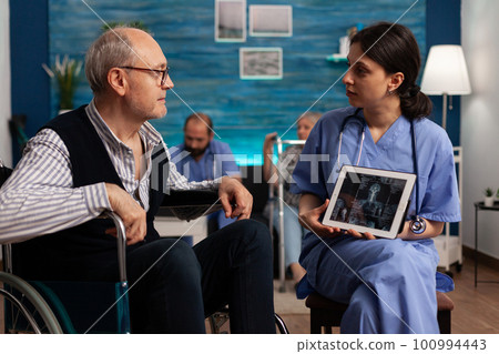 Female nurse explaining medical radiography with a tablet to an elderly patient with disability. Retired man in wheelchair at physical examination appointment at health center. Female nurse explaining medical radiography with a tablet to an elderly patient with disability. Retired man in wheelchair at physical examination appointment at health center. 100994443