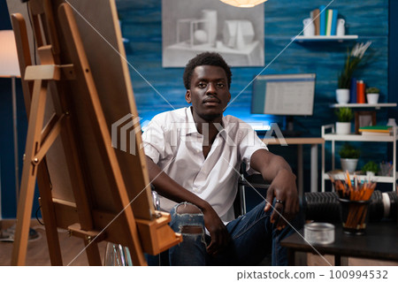 Plastic arts student using wheelchair, drawing on easel. African american artist living with chronical condition creating masterpiece in home art studio. Cartoonist in modern atelier looking at camera Plastic arts student using wheelchair, drawing on easel. African american artist living with chronical condition creating masterpiece in home art studio. Cartoonist in modern atelier looking at camera 100994532