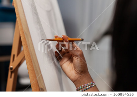 Close up of hand of unrecognizable african american female artist making strokes with pencil on canvas easel in drawing workshop. Unidentified creative person finishing artwork at studio. Close up of hand of unrecognizable african american female artist making strokes with pencil on canvas easel in drawing workshop. Unidentified creative person finishing artwork at studio. 100994536