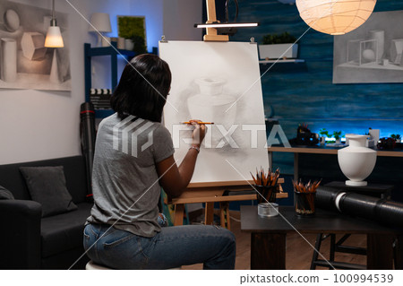 Young african american woman with artistic abilities sketching pot at workplace with creative tools. Professional female artist finishing pencil drawing of vase at home made art studio. 100994539