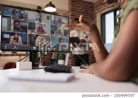 African american woman gesturing in internet meeting with colleagues while roommate is working on laptop in home living room. Startup employee in internet call with diverse team using pc and webcam. 100994597
