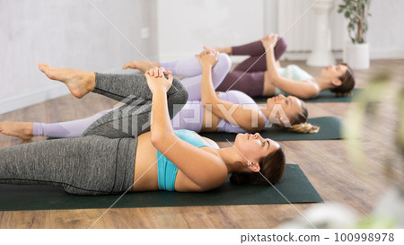 Class of young sporty women stretching gluteal muscle while lying on back during yoga Pilates workout on mats in gym studio Class of young sporty women stretching gluteal muscle while lying on back during yoga Pilates workout on mats in gym studio 100998978