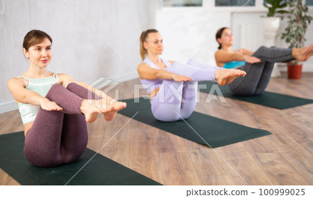 Calm sportive women performing yoga boat pose on mats Paripurna Navasana during group training in fitness studio Calm sportive women performing yoga boat pose on mats Paripurna Navasana during group training in fitness studio 100999025