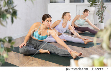 Diligent women practicing hand to toe pose of yoga in light fitness room Diligent women practicing hand to toe pose of yoga in light fitness room 100999196