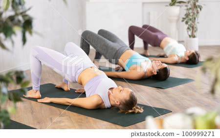 Sporty young women practicing Bridge Pose Setu Bandha Sarvangasana during group yoga workout on retreat 100999271