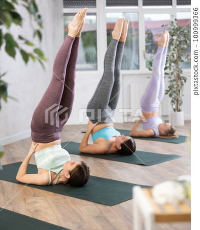 Active fit girls in sportswear, keeping bodies in vertical position and resting on shoulders during Sarvangasana in yoga class 100999366