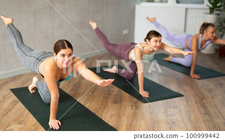 Diligent women practicing balancing table pose of yoga in light fitness room Diligent women practicing balancing table pose of yoga in light fitness room 100999442