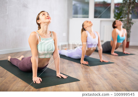 Diligent women practicing cobra pose of yoga in light fitness room 100999545