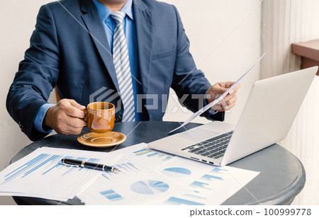 Businessman hand holding coffee cup, analysis the graph with laptop at the home office for setting challenging business goals And planning to achieve the new target 100999778