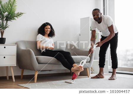 Smiling woman working on laptop while man vacuuming carpet Smiling woman working on laptop while man vacuuming carpet 101004457