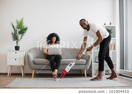 Husband cleaning with vacuum while wife working on laptop 101004459