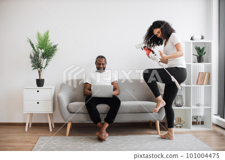 Woman dancing with cordless vacuum while man using laptop Woman dancing with cordless vacuum while man using laptop 101004475