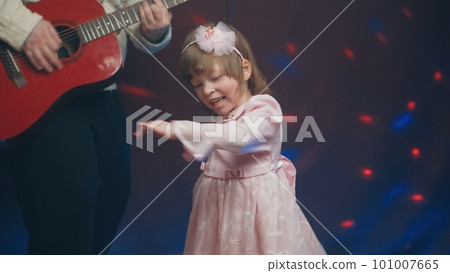 Little girl on stage in pink vintage dress, she dances, her father plays acoustic guitar. Color music is shining. Performance on stage. Early development of children Little girl on stage in pink vintage dress, she dances, her father plays acoustic guitar. Color music is shining. Performance on stage. Early development of children 101007665
