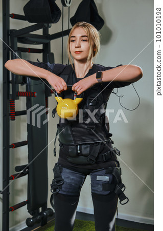 Beautiful blonde woman wearing in sensors and belts, fastens with velcro is practicing EMS fitness in a gym, building up muscles with a kettlebell. Beautiful blonde woman wearing in sensors and belts, fastens with velcro is practicing EMS fitness in a gym, building up muscles with a kettlebell. 101010198