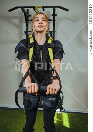 Beautiful blonde woman wearing in sensors and belts, fastens with velcro is practicing EMS fitness in a gym performing traction exercises. Beautiful blonde woman wearing in sensors and belts, fastens with velcro is practicing EMS fitness in a gym performing traction exercises. 101010210
