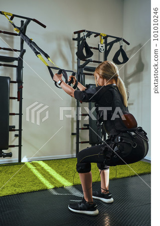 Beautiful blonde woman wearing in sensors and belts, fastens with velcro is practicing EMS fitness in a gym performing traction exercises. Beautiful blonde woman wearing in sensors and belts, fastens with velcro is practicing EMS fitness in a gym performing traction exercises. 101010246