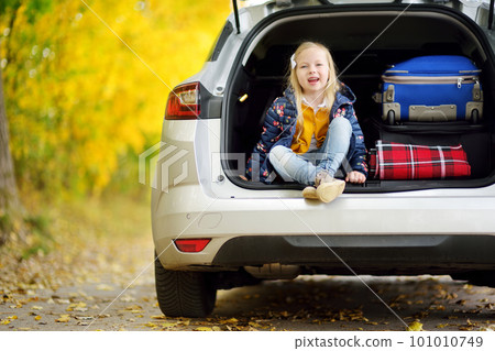 Adorable girl sitting ain a car trunk ready to go on vacations with her parents. Child looking forward for a road trip or travel. Autumn break at school. 101010749