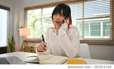 Charming young Asian woman freelancer having phone conversation and making note on notebook Charming young Asian woman freelancer having phone conversation and making note on notebook 101011520
