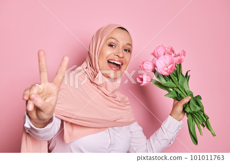 Gorgeous Arab Muslim woman wearing pink hijab, holding a bouquet of tulips, showing peace hand sign, smiling a beautiful toothy smile, looking at web camera while making selfie over pink background 101011763