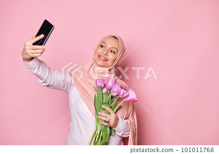 Attractive Muslim pregnant woman in pink hijab, holding a bouquet of purple tulips, smiling a toothy smile, looking at web camera while making selfie on her smartphone, isolated over pink background Attractive Muslim pregnant woman in pink hijab, holding a bouquet of purple tulips, smiling a toothy smile, looking at web camera while making selfie on her smartphone, isolated over pink background 101011768
