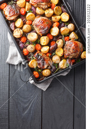 Delicious homemade chicken thighs baked with potatoes, garlic, onions and tomatoes in balsamic vinegar close-up on a black baking sheet. Vertical top view 101012222