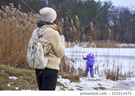 Mother watching a child playing on the shore of a frozen lake in a winter park 101012471