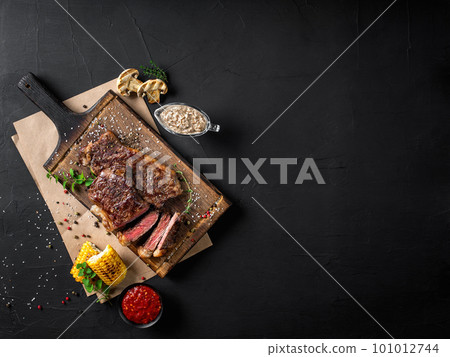 Fried steaks with herbs and spices on wooden board, parchment, grilled garlic, corn, two sauces on black background. Close-up. Top view. 101012744