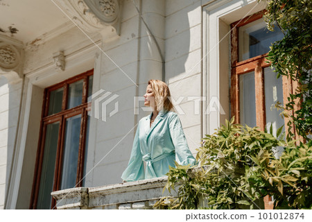 A woman on a balcony in a long blue dress stands near a low granite stone fence against the background of an old stone house A woman on a balcony in a long blue dress stands near a low granite stone fence against the background of an old stone house 101012874