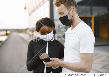 African american fitness model and caucasian man looking at the phone while training outdoors. Woman wearing black sportswear. Couple wearing a face mask. 101013844