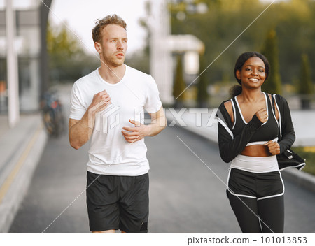 African american fitness model and caucasian man jogging outdoors. Woman wearing black sportswear. Man wearing white t-shirt. 101013853