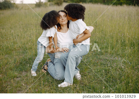 Photo of caucasian mother and two her african american daughters embracing together outdoors. Girl shas black curly hair. Mother and daughers wearing white t-shirts. Photo of caucasian mother and two her african american daughters embracing together outdoors. Girl shas black curly hair. Mother and daughers wearing white t-shirts. 101013929