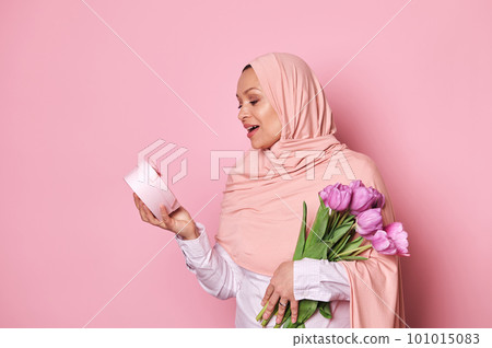 Surprised Muslim pretty woman in pink hijab, holding a bouquet of tulips, expressing amazement while getting a happy present in a pink gift box for festive occasion, isolated over pink background 101015083