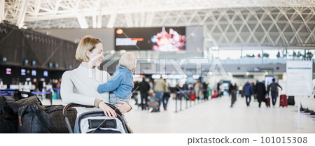 Motherat travelling with his infant baby boy child, walking, pushing baby stroller and luggage cart at airport terminal station. Travel with child concept 101015308