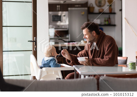 Father wearing bathrope spoon feeding hir infant baby boy child sitting in high chair at the dining table in kitchen at home in the morning. Father wearing bathrope spoon feeding hir infant baby boy child sitting in high chair at the dining table in kitchen at home in the morning. 101015312