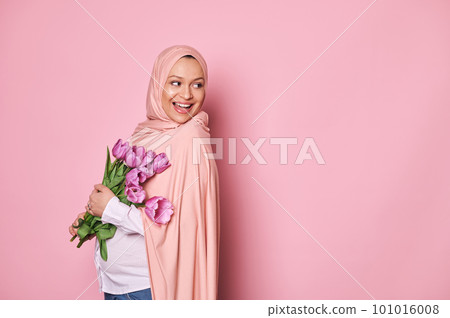 Gorgeous Middle-Eastern Muslim woman dressed in pink hijab, smiling looking through her shoulder at a copy space on isolated pink background, expressing happiness, posing with a cute bouquet of tulips 101016008
