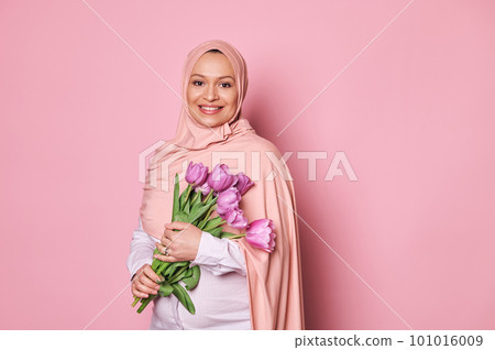 Confident portrait of a beautiful young Muslim pregnant wearing a pink hijab, holding a bouquet of purple tulips, smiling looking at camera, isolated on pink background. Happy Mother's Day concept Confident portrait of a beautiful young Muslim pregnant wearing a pink hijab, holding a bouquet of purple tulips, smiling looking at camera, isolated on pink background. Happy Mother's Day concept 101016009