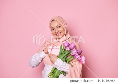Positive Middle-Eastern Muslim woman in pink hijab with Mother's Day, birthday present or for any festive occasion and bouquet of purple tulips, smiling looking at camera, on isolated pink background 101016862