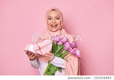Cheerful Arab lady in pink hijab, holding a heart shaped gift box and tulips, smiling to camera. Positive amazed Muslim woman with Mother's Day or birthday present posing over pink studio background Cheerful Arab lady in pink hijab, holding a heart shaped gift box and tulips, smiling to camera. Positive amazed Muslim woman with Mother's Day or birthday present posing over pink studio background 101016863