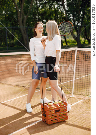 Female friends with racquets and box of balls on the tennis court. Summer, outside court. 101017029