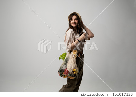 Concept of no plastic. Zero waste, plastic free. Sustainable lifestyle. Happy woman in a beige t-shirt and a hat holding reusable cotton shopping bag with organic groceries, bread and greens. Concept of no plastic. Zero waste, plastic free. Sustainable lifestyle. Happy woman in a beige t-shirt and a hat holding reusable cotton shopping bag with organic groceries, bread and greens. 101017136