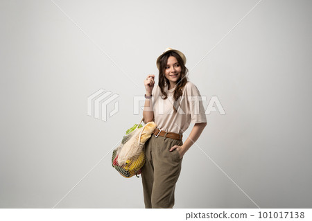 Concept of no plastic. Zero waste, plastic free. Sustainable lifestyle. Happy woman in a beige t-shirt and a hat holding reusable cotton shopping bag with organic groceries, bread and greens. Concept of no plastic. Zero waste, plastic free. Sustainable lifestyle. Happy woman in a beige t-shirt and a hat holding reusable cotton shopping bag with organic groceries, bread and greens. 101017138
