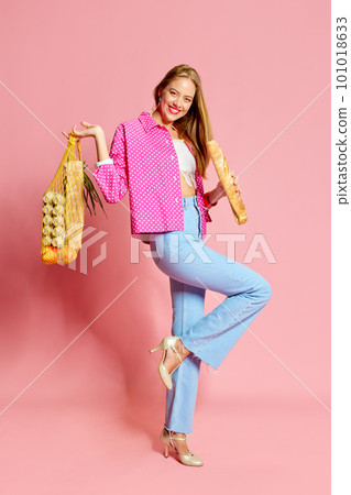 Cheerful young woman wearing jeans, heels holding purchases and baguette over pink background Cheerful young woman wearing jeans, heels holding purchases and baguette over pink background 101018633