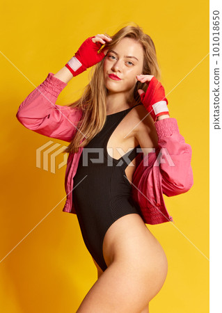 Portrait of adorable woman wearing bodysuit and bandages for boxing touching hair and looking at camera over yellow background 101018650