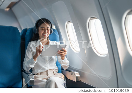Young Asian business woman or female passenger wearing wireless headphone and working with tablet during the flight 101019089