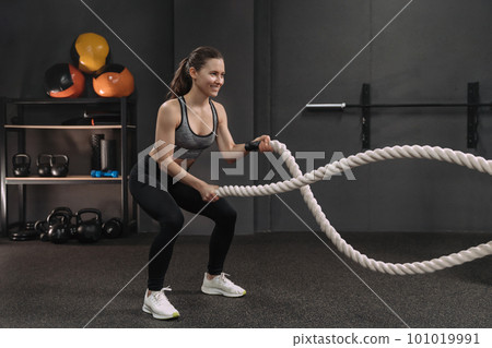 Muscular young smiling woman working out with battle ropes at dark gym, 101019991