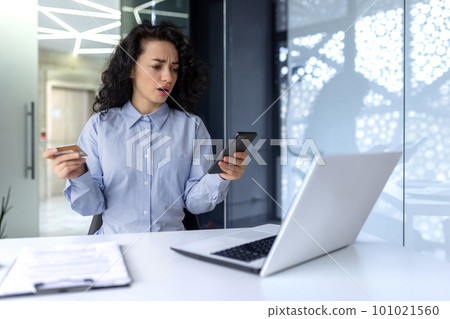 Credit card problems. A young Arab woman is sitting at the desk in the office and looking worriedly at the phone. Unsuccessful shopping, blocked channel. Credit card problems. A young Arab woman is sitting at the desk in the office and looking worriedly at the phone. Unsuccessful shopping, blocked channel. 101021560